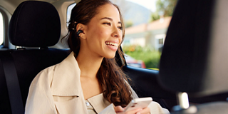 Kvinde, som bærer Bose Ultra åbne øretelefoner, mens hun sidder på bagsædet i en bil og lytter til musik fra sin telefon