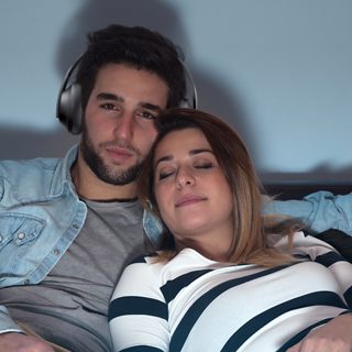 Hombre viendo televisión utilizando unos audífonos Bose Noise Cancelling Headphones 700 mientras una mujer duerme
