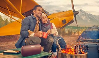 A couple sitting by a fire listening to music from a Bose SoundLink Flex Bluetooth Speaker with an orange Carbon Cub SS behind them