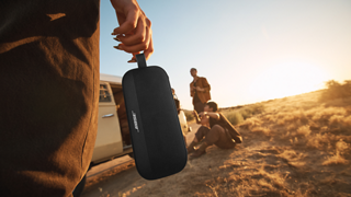 Woman using the utility strap to carry a Bose SoundLink Flex Portable Speaker (2nd Gen) to a group of friends sitting next to a van.